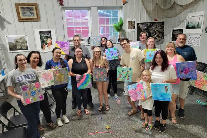 Group of people smiling and holding colorful paintings in an art studio.