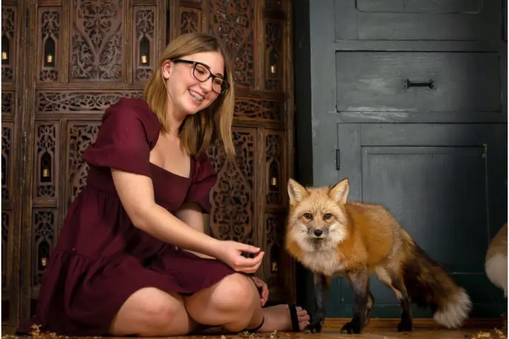 Woman in burgundy dress sitting on floor with a fox indoors.